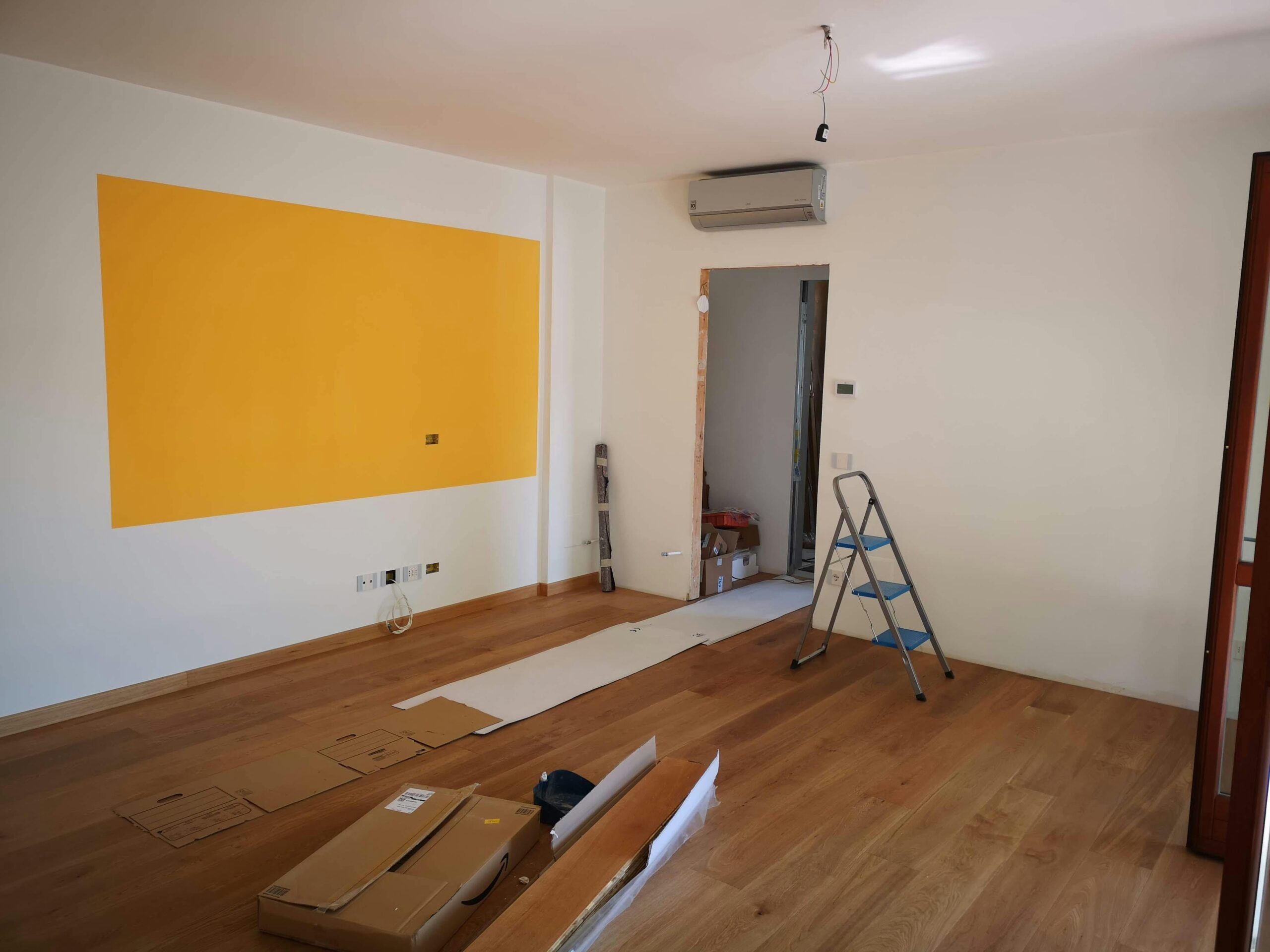 A bright, empty room under renovation with ladder, tools, and wooden flooring.