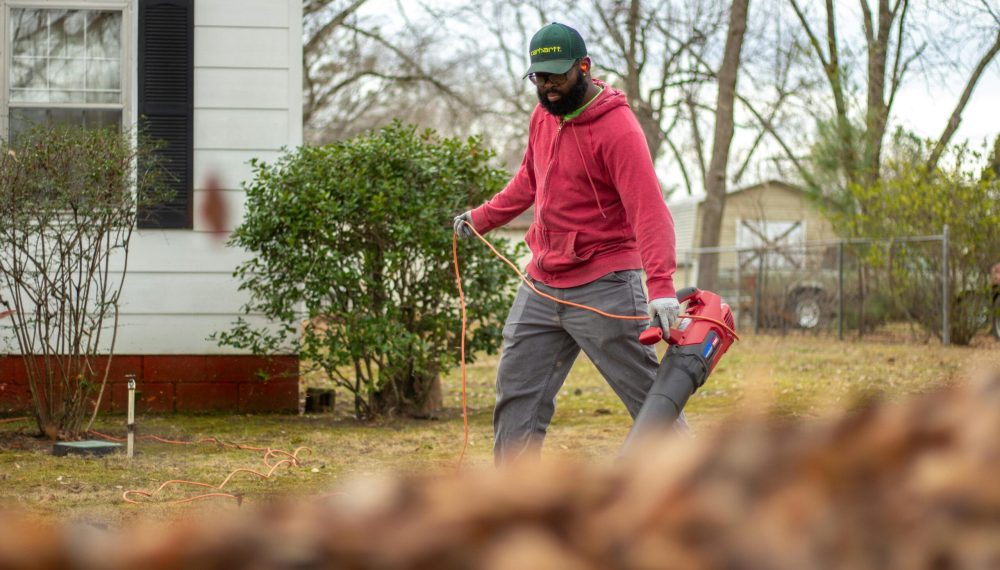 A man in a red jacket operates a leaf blower in an autumn backyard, clearing leaves.